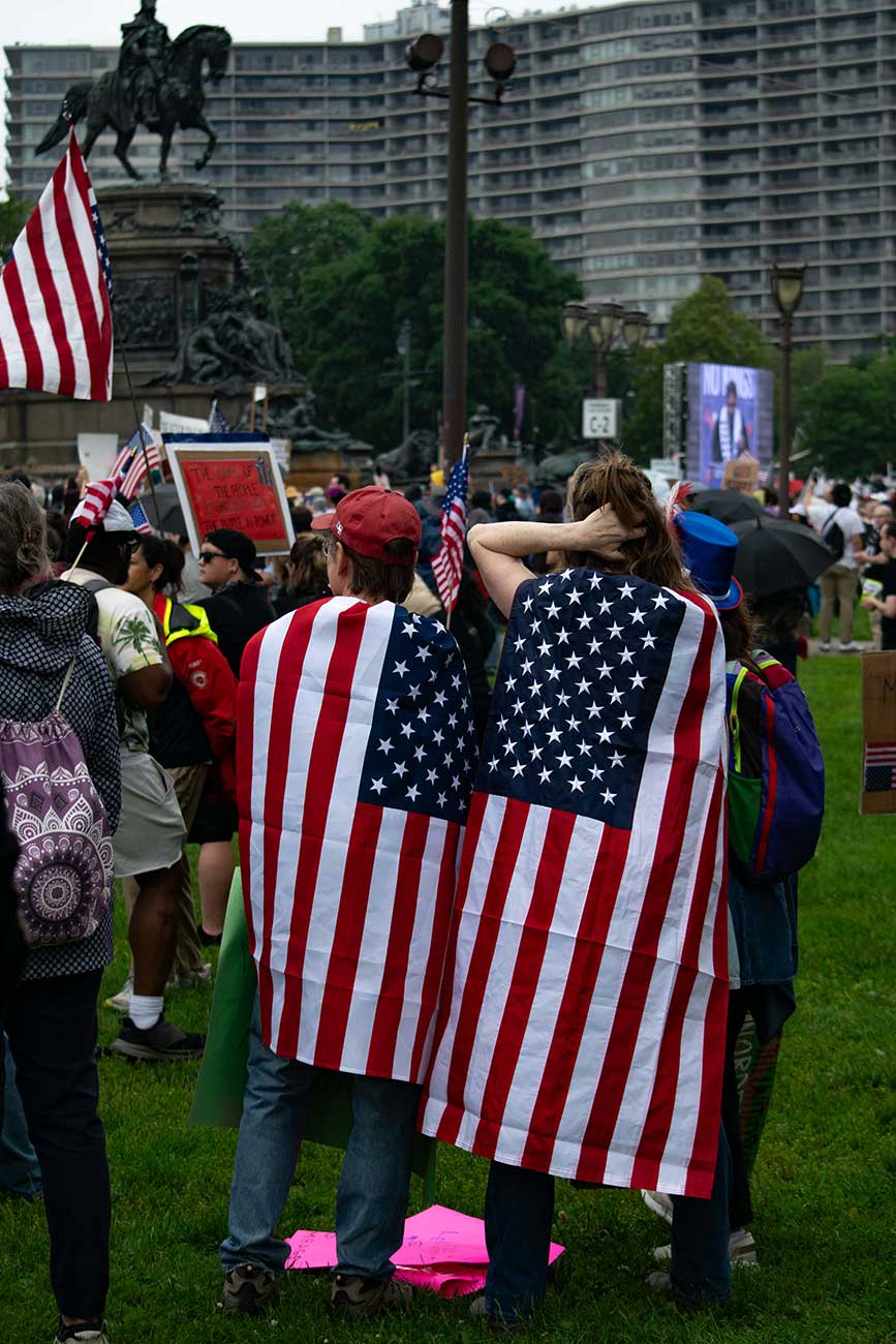 Two protesters wear American flags as capes as they listen to speakers at the No Kings Day Protest in front of the Philadelphia Art Museum